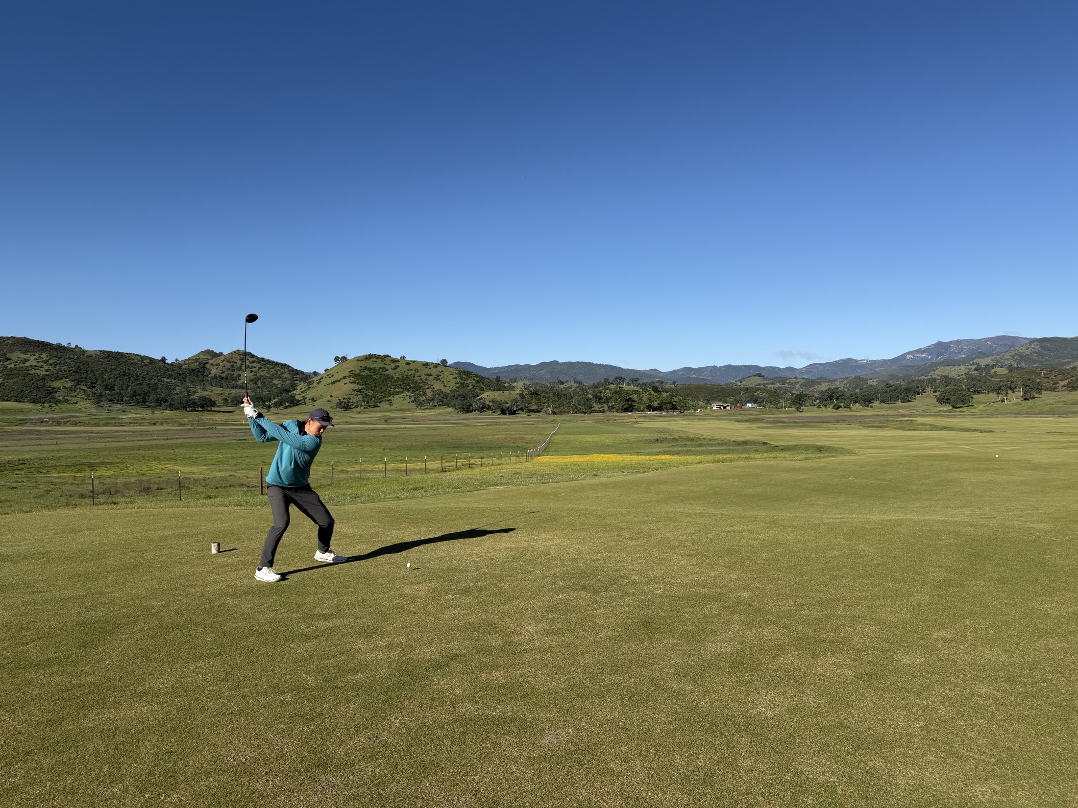 Charles mid-swing on a sunlit tee, with rolling hills beyond the fairway.